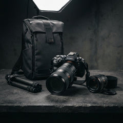 Professional photography gear display featuring a Sony mirrorless camera with a large lens, a secondary camera lens, a folded carbon fiber tripod, and a dark gray camera backpack on a textured stone surface under studio lighting.