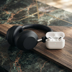 Matte black Sony over-ear headphones and white wireless earbuds in an open charging case displayed on a wooden board over a green marble surface.