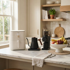 Modern kitchen setup featuring a cream-colored Cosori air fryer, a sleek black electric gooseneck kettle, a black coffee grinder, and a ceramic pedestal bowl filled with fresh fruit on a white marble countertop.