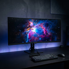 Large ultra-wide curved desktop monitor labeled AETHERIS displaying a vibrant cosmic space nebula wallpaper, paired with a mechanical keyboard and wireless mouse on a carbon fiber texture desk with blue ambient backlighting.